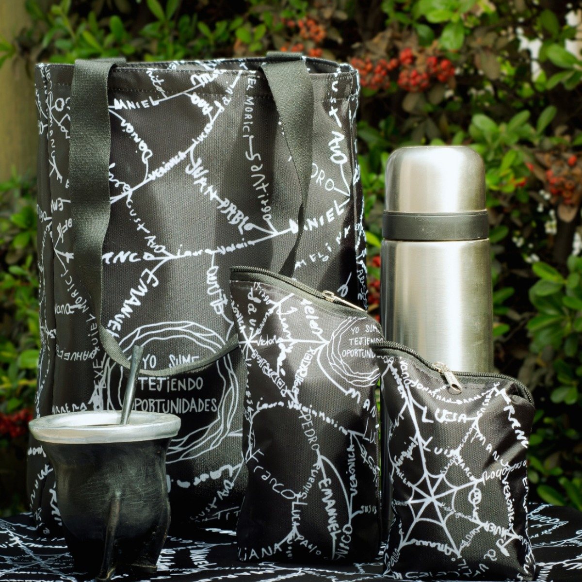 Set matero negro con la trama de nombres en blanco: bolso, yerbero y azucarero, junto a un termo y un mate, al aire libre.